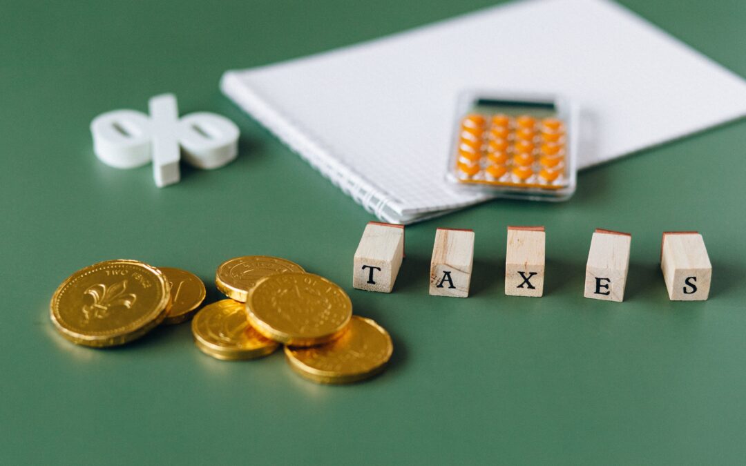 A calculator on top of a notepad, a percentage symbol, taxes spelled out of wooden blocks, and gold coins kept on a green surface.