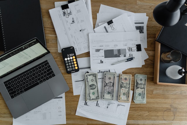 A laptop, stacks of dollars, a calculator, and papers on a wooden table.